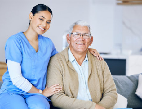 a caregiver sitting on a couch with an elderly man, smiling at the camera